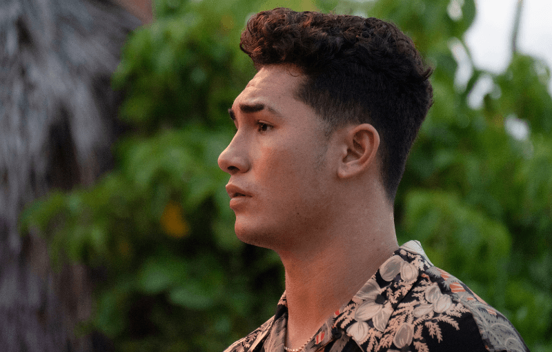 A young man with short curly hair looks to the side against a backdrop of lush green foliage. He is wearing a patterned shirt and appears thoughtful, with the image captured in natural light.