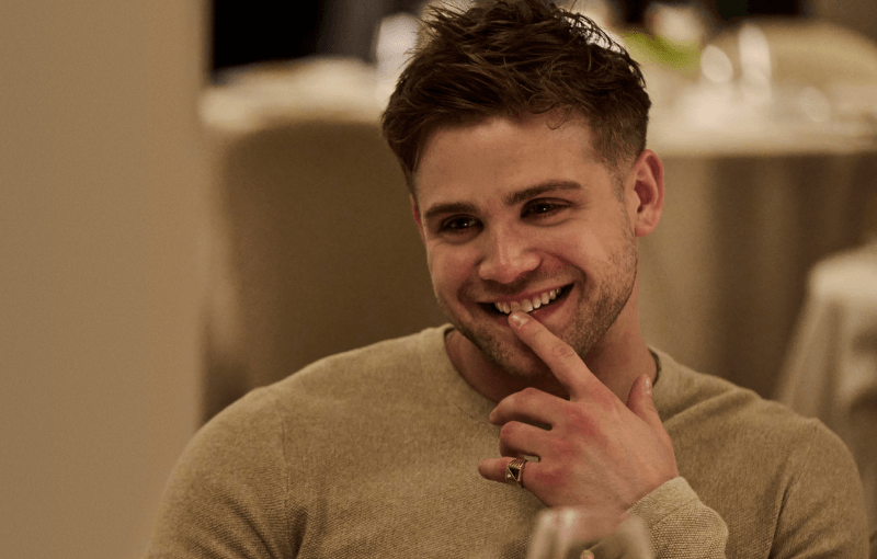 A man with short hair and wearing a beige sweater is smiling and gently touching his lips with his fingers. He is sitting in what appears to be a restaurant, suggested by the blurred background of tables and chairs.