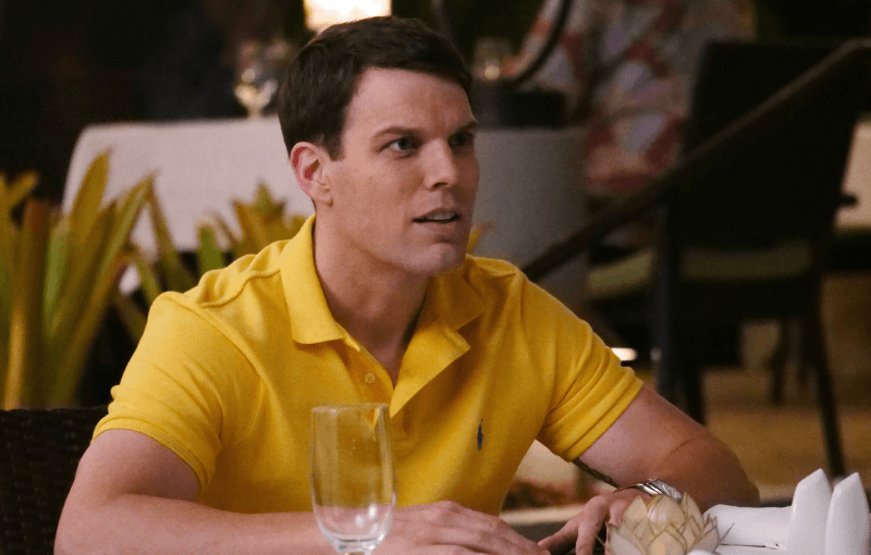 A man in a yellow polo shirt sits at a table in a restaurant, looking to the side. There are empty glasses and folded napkins on the table. The background is softly blurred, with plants and dim lighting.