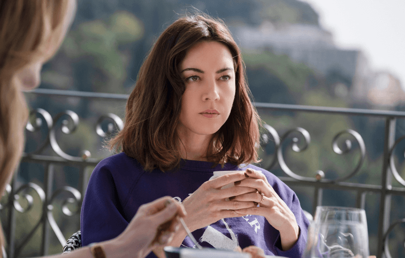 Woman with shoulder-length hair holds a cup, sitting outdoors at a table. She wears a purple top and looks pensive. The background shows a blurred view of greenery and buildings, with a wrought iron railing.