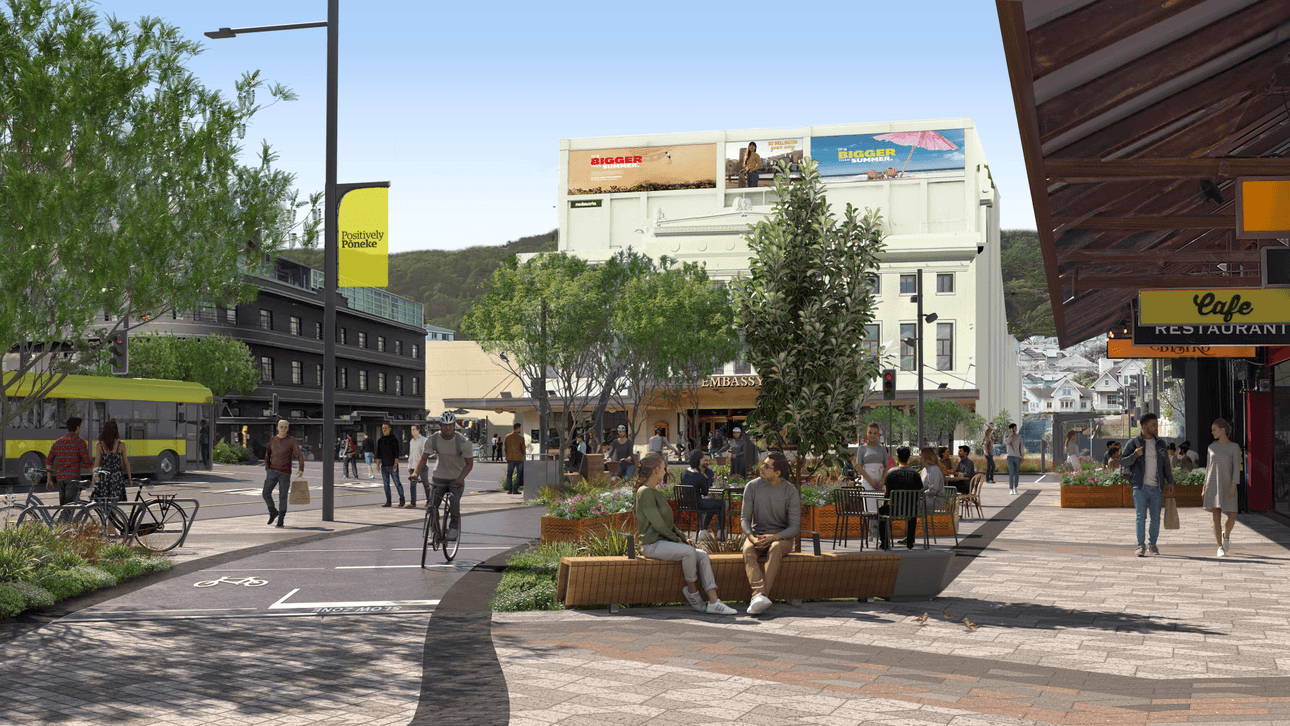 A bustling urban street scene with people walking, biking, and sitting on benches. Trees and planters line the sidewalk. Shops and cafes are visible, with a clear sky above.