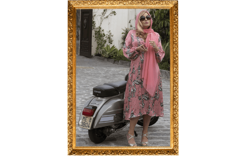 A person in a pink dress and sunglasses stands beside a vintage silver scooter on a cobblestone street. They are framed by a gold border, with a pink headscarf and matching shoes. Walls with greenery are in the background.