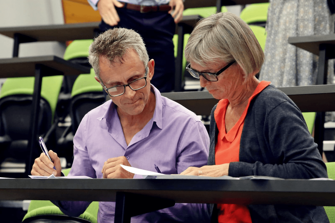 Two people sitting at a desk in a lecture hall, both wearing glasses. The person on the left is in a purple shirt, holding a pen and looking at papers. The person on the right is in a red top and grey cardigan, also holding papers, with green seats in the background.