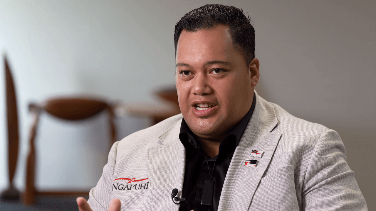 A person wearing a light-colored blazer with "Ngapuhi" embroidered on it speaks and gestures with their hand. The background is blurred with wooden chairs visible.