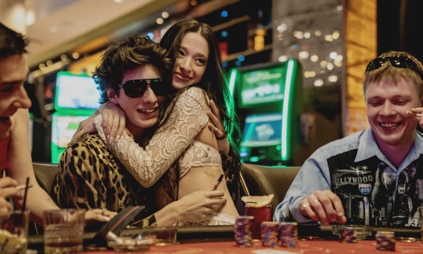 A lively scene at a casino from Anora. Four people are sitting around a poker table. A woman (Ani) in a sparkling dress hugs a man in sunglasses (Vanya). Poker chips and cards are on the table, while another man smiles, holding cards. Bright lights are visible in the background.