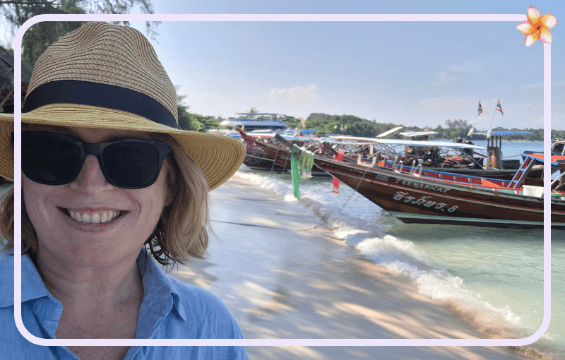 A person in sunglasses and a wide-brimmed hat smiles at the camera on a beach. Long-tail boats are moored in the shallow water behind them, and there are flags on the boats. The sky is clear and sunny. There's a flower graphic in the top corner.