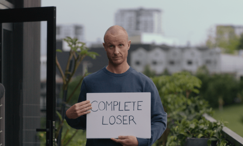 Jono Pryor stands outside holding a sign that says "COMPLETE LOSER" with a neutral expression. The background includes a blurred outdoor setting with greenery and buildings.