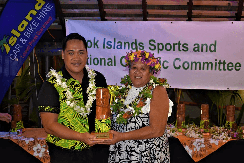 A man and a woman, both adorned with floral garlands, stand together smiling. The man holds a trophy. A banner behind them reads "Cook Islands Sports and National Olympic Committee." Tables with trophies and floral decorations are in the background.