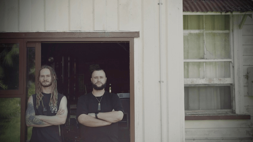 Two brothers stand with arms crossed in front of a house