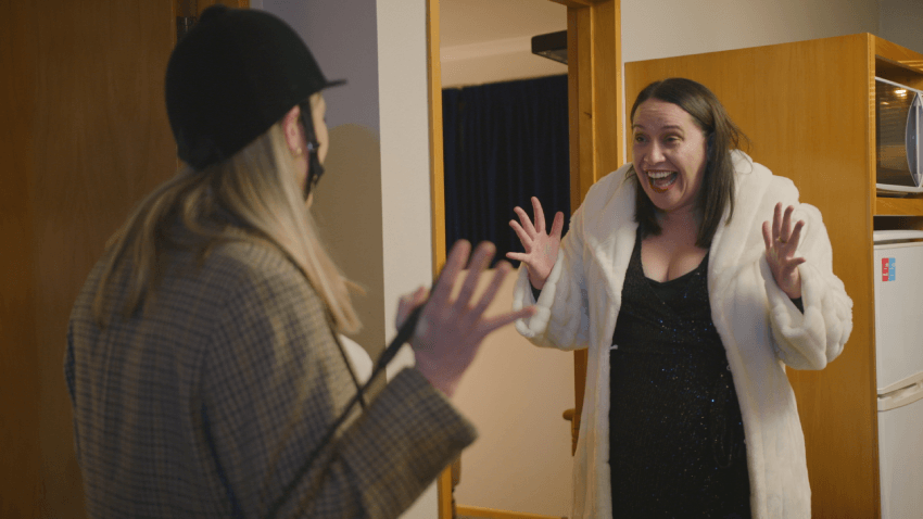 Brynley Stent and Kura Forrester are interacting warmly in a room. One wears a stylish coat, a hat, and long hair, while the other, with short hair, wears a shimmering black dress and a white fur coat. They appear surprised and happy, with arms raised in excitement.