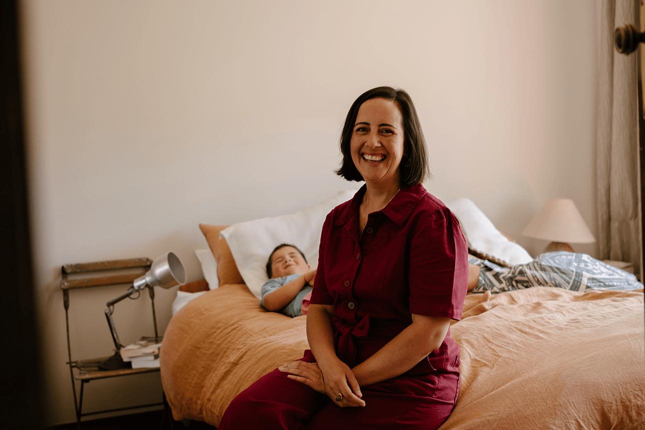 woman sitting on a bed in red jumpsuit - child lying down behind her