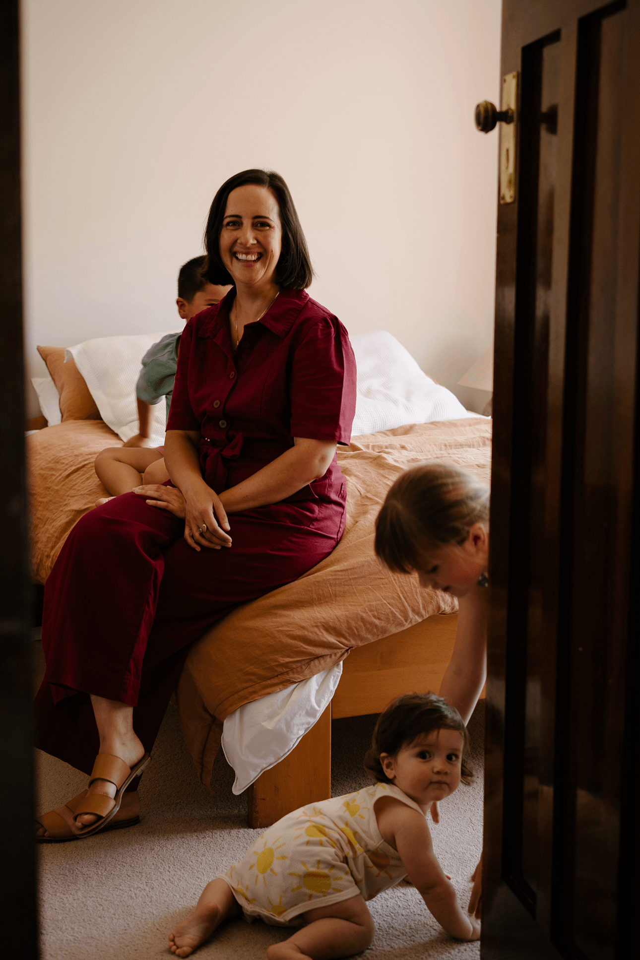 woman in red jumpsuit on a bed with three children playing around her