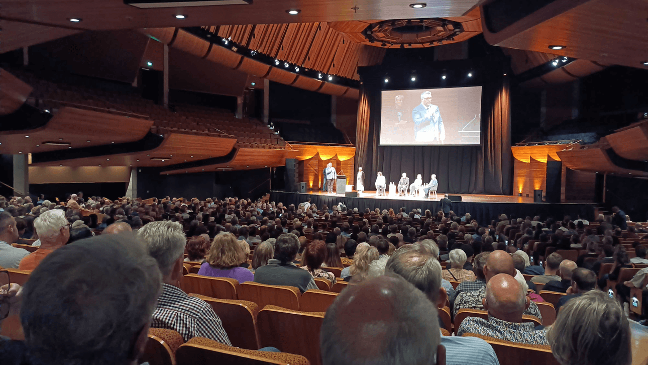 A photo taken at the back of a crowd of people, many with grey hair, watching four panellists on stage