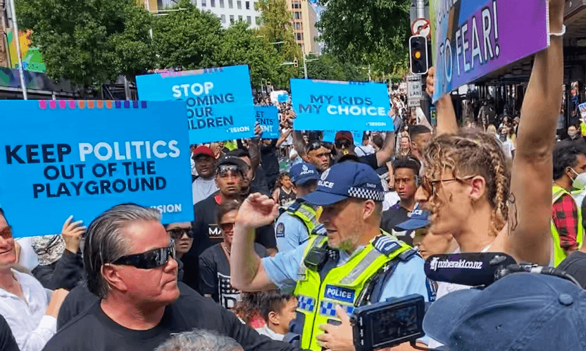 A protest with people holding signs such as "Keep politics out of the playground" and "My kids, my choice." A police officer talks to Brian Tamaki. The crowd is diverse, with trees and buildings in the background.