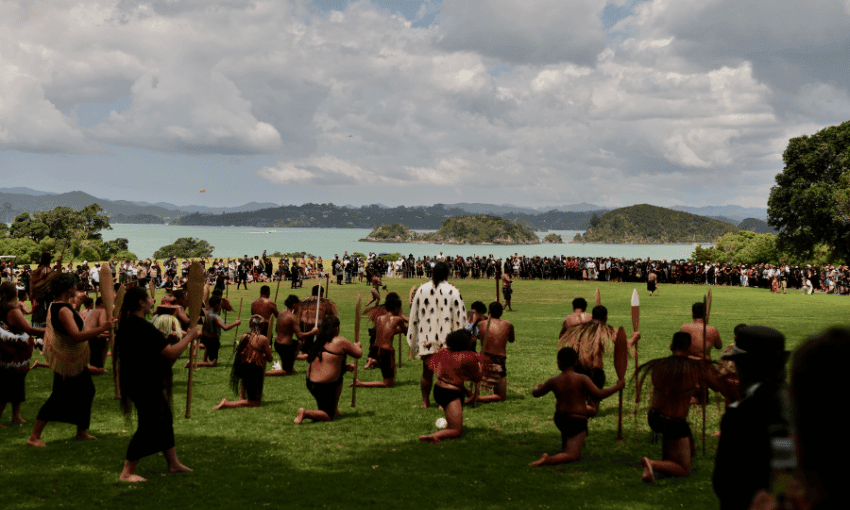 A traditional Māori ceremony is taking place on a lush green field. Participants, some wearing cultural attire and holding spears, are kneeling. A large gathering observes against a backdrop of water and islands under a cloudy sky.