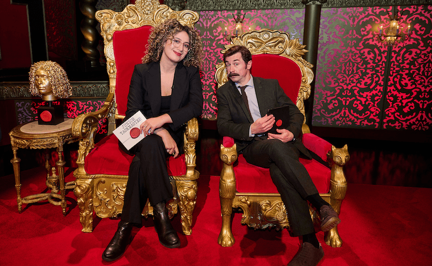 A woman with curly hair wearing a black suit sits in a red velvet Taskmaster throne holding an envelope