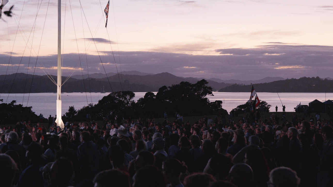 A darkened shot at sunrise, with a crowd of people gathered on the upper Treaty Grounds at Waitangi, watching a big screen, not pictured