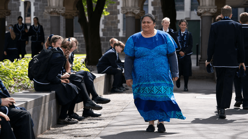 A Samoan woman in a vibrant blue patterned dress walks through the uniformed school yard