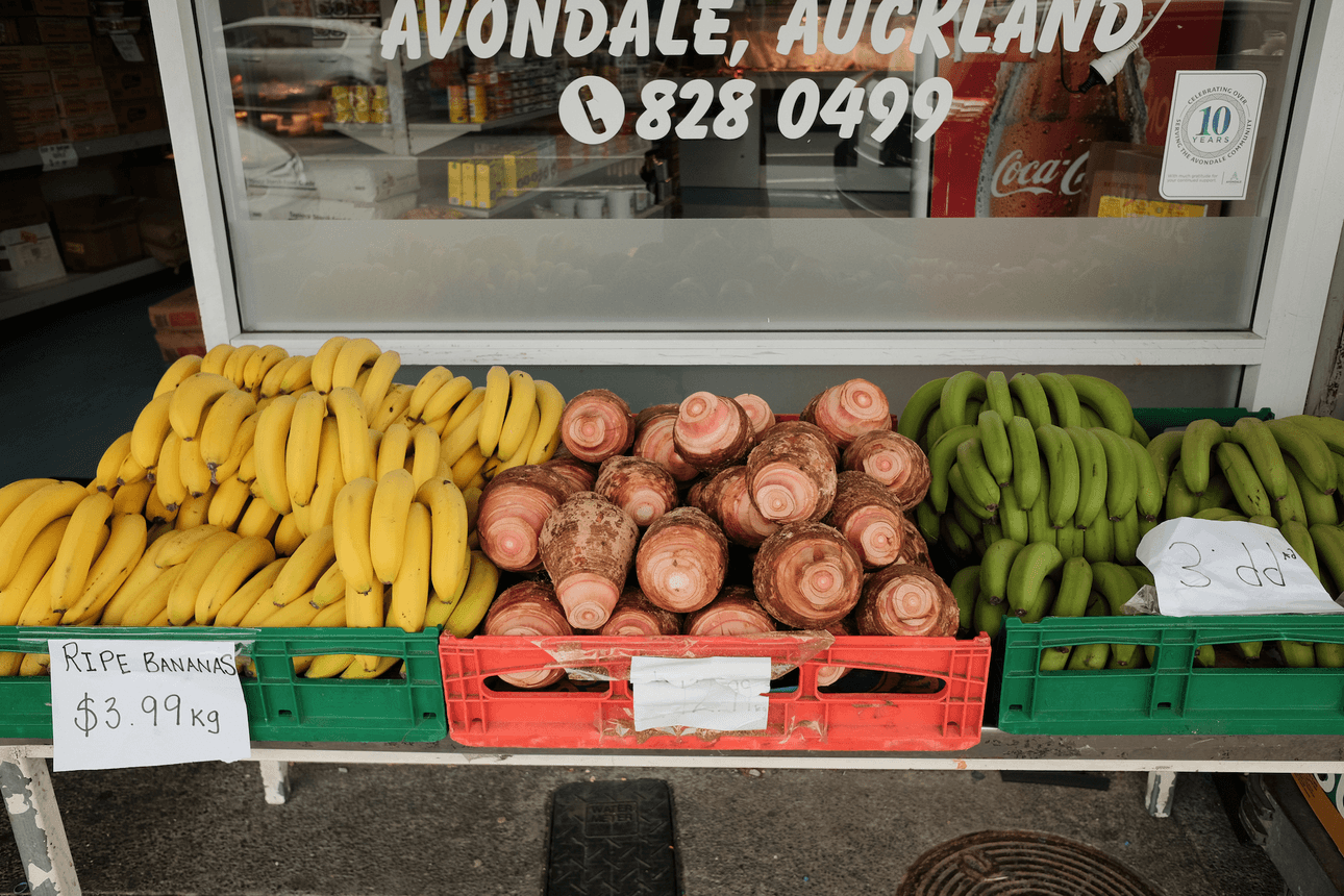 Yellow bananas, raw taro and green bananas for sale on a table. Behind the table is a shop window with partial shop signage that reads "avondale, auckland"