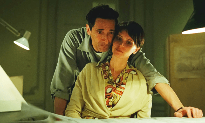 A man and woman are sitting closely together in a dimly lit room, looking pensive. The woman wears a patterned shirt and cream cardigan, while the man is dressed in a blue shirt. Papers and a desk lamp are visible in the foreground.