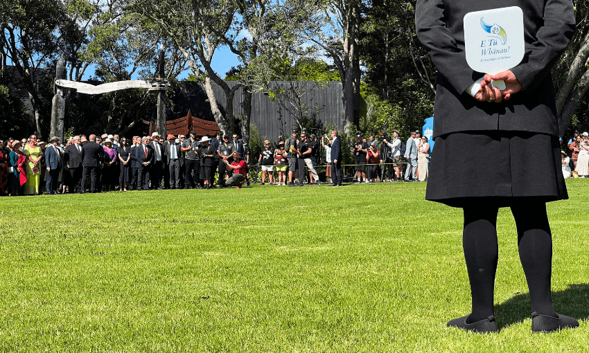 Politicians gather on the lawn at the Upper Treaty Grounds at Waitangi. In the foreground, a Māori warden dressed in black with a white fan behind their back watches the crowd.