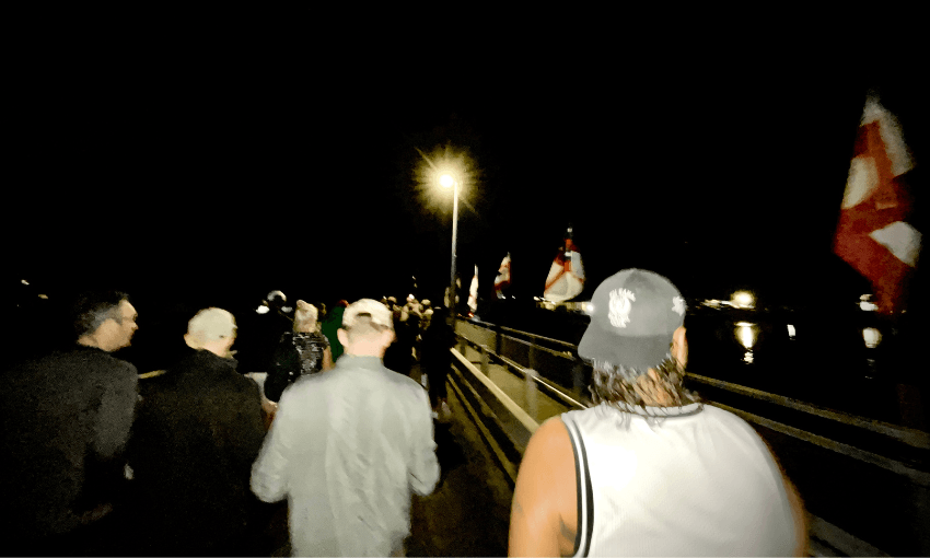 A group of people walk along the dimly lit bridge at Waitangi at dawn. Flags are visible on the right side of the bridge. The scene is illuminated by a bright streetlight in the distance.