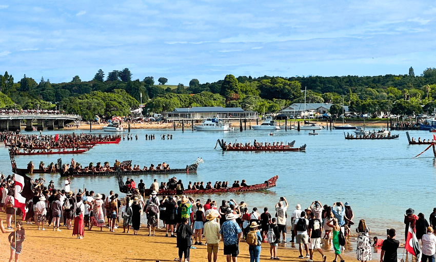 A large crowd gathers on a sandy beach to watch waka in a scenic bay. The backdrop includes green hills, trees, and a few buildings near the shoreline under a partly cloudy sky.