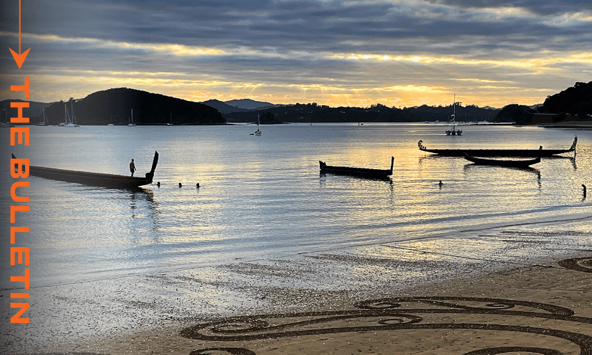 Waka at Waitangi, February 6, 2025 (Photo: Anna Rawhiti-Connell) Serene beach scene at dawn at Waitangi with several waka silhouetted on calm water. Gentle waves lap the shore, which features intricate sand art in the foreground. Text on the left reads "The Bulletin".