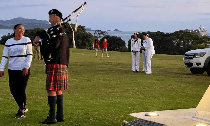 A person in a kilt with bagpipes on a grassy field, with a smiling person walking nearby. In the background, two individuals in white naval uniforms stand near a vehicle, and there are trees and a distant ocean view.