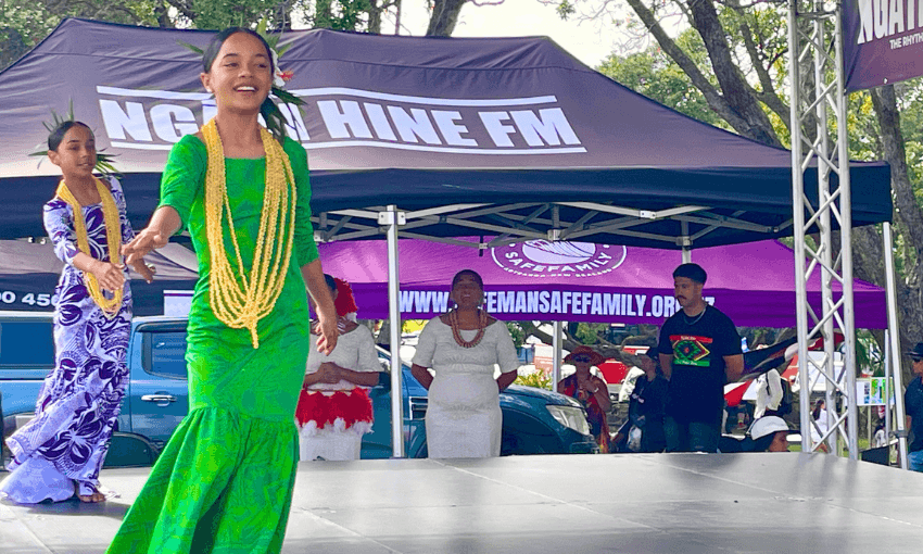 Young people perform a dance on an outdoor stage. A girl in a green dress leads. A purple tent in the background has "NGĀTI HINE FM" on a banner. Trees and people are visible in the background.