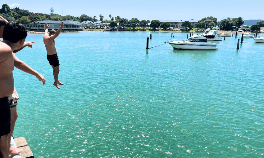 A group of people on the bridge at Waitangi one in mid-air jumping into the water. Boats and a distant shoreline with trees and buildings are visible under a clear blue sky.