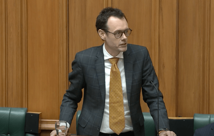 A person wearing glasses, a dark checkered suit, a white shirt, and a mustard tie stands at a lectern, holding a glass of water. The background features wooden paneling.