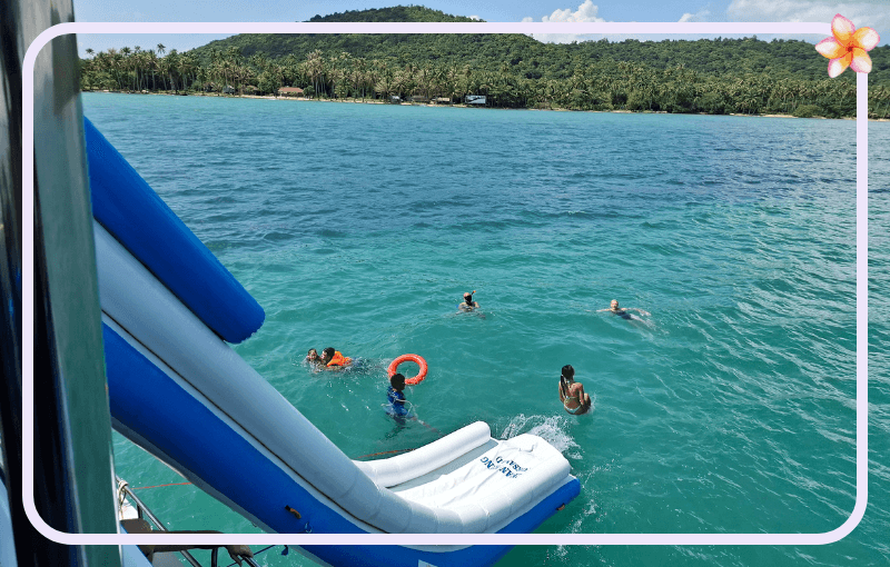 People swim in clear turquoise waters near a blue and white inflatable slide extending from a boat. Green hills and palm trees are visible in the background. A red circular float is being used by one of the swimmers.