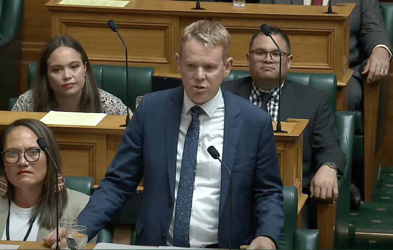 A man in a blue suit and tie speaks at a wooden lectern in a government chamber. Seated behind him are three people, two men and a woman, listening attentively. Microphones are visible at their desks.