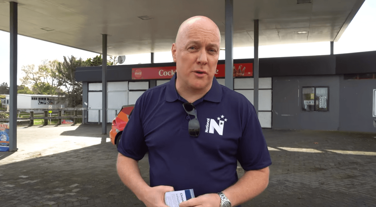 Chris Luxon in a blue National polo stands in front of a former Cockle Bay dairy. The sign on the dairy is cut off by his head and rather unfortunately just reads "cock"