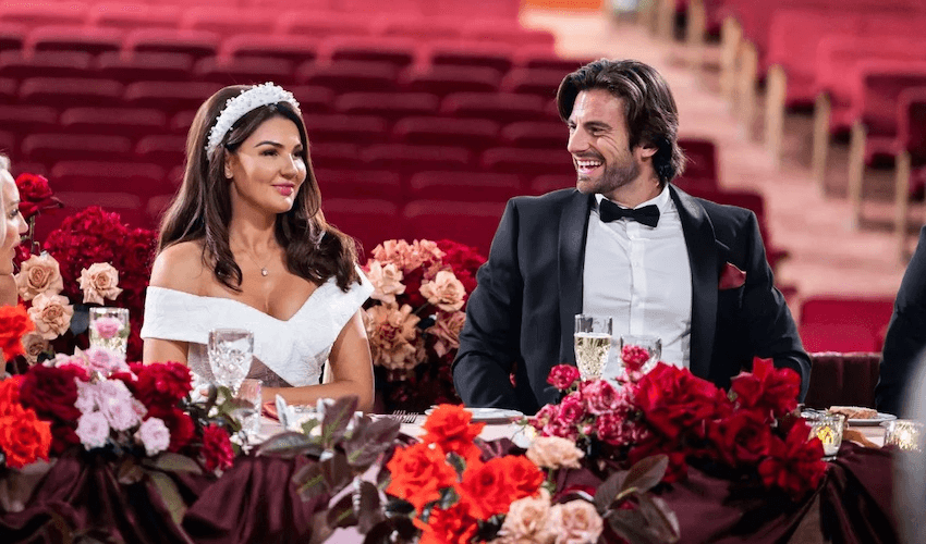 Lauren sits in a white wedding dress at the head table with Eliot in his suit. They are smiling, for now