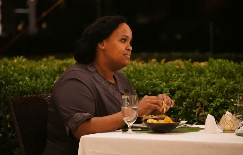 A person seated at an outdoor table, holding food and looking to the side. The table has a water glass and a bowl of snacks. In the background, green bushes are visible. They are wearing a dark shirt and it's evening.