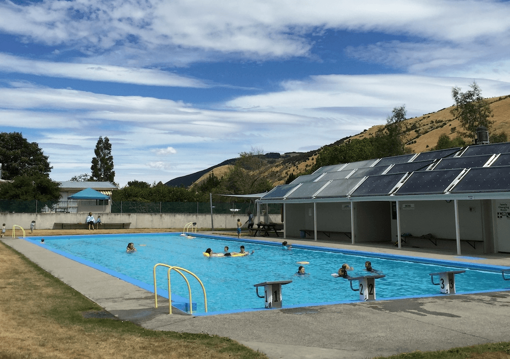 a cloud flecked sky and the glimpse of dark yellow summery central Otago hills. In the foreground, a pool with a yellow ladder and phalanx of solar panels is filled with people, mostly kids, swimming