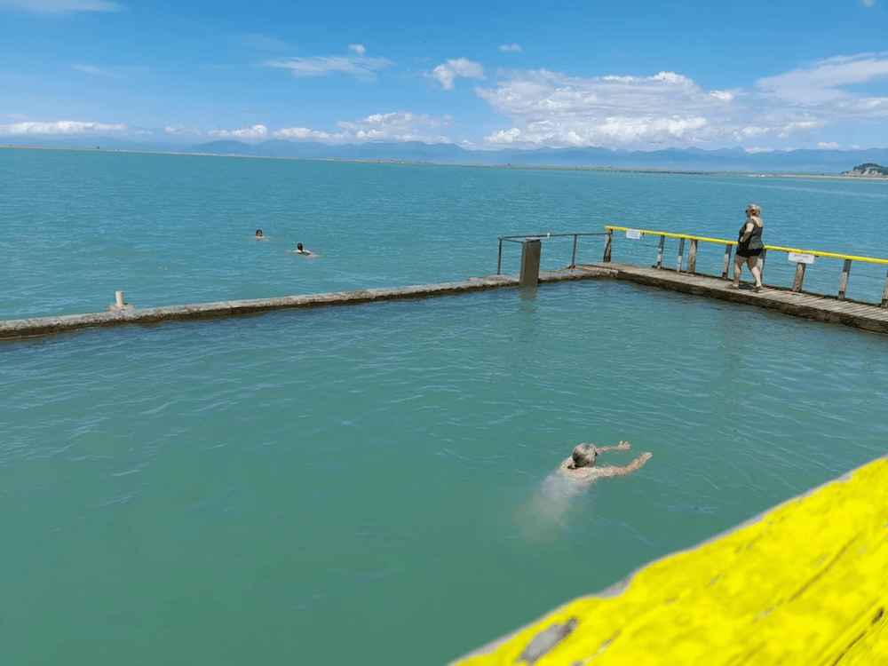 a slightly cloudy green pool, with a line of concrete separating it from the ocean beyond. someone is swimming in the centre of the picture with their arms out