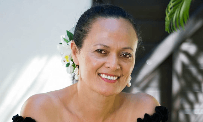 A person with a flower in their hair smiles warmly, wearing off-shoulder black attire and pearl earrings. They're standing outdoors, with greenery and natural light in the background.