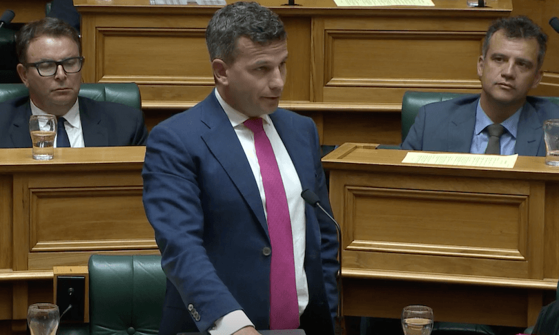 A man in a suit and pink tie speaks at a podium in a formal setting, possibly a government or legislative chamber. People in suits are seated nearby, listening attentively. Wooden desks and glass water bottles are visible.