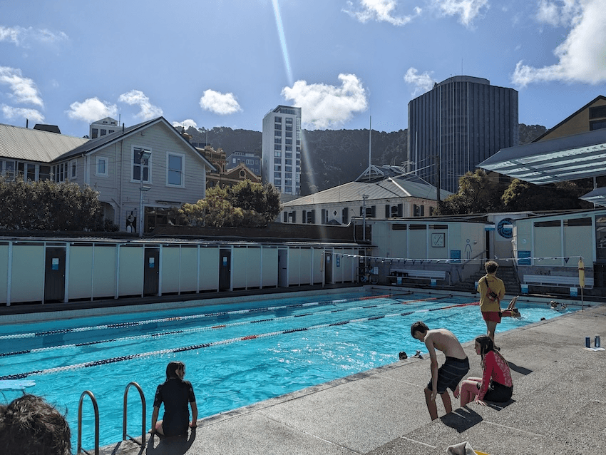 Throndon pools, with kids lurking on the concrete bleaches and the water illuminated in bright ultramarine with a stripe of sunlight slicing through the buildings and hills visible beyond