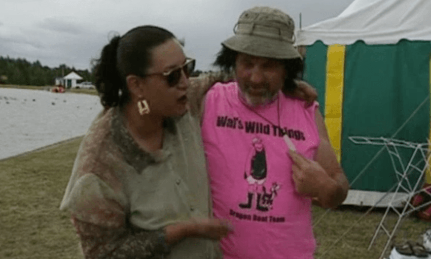 Georgina Beyer, dressed glamorously in a silk shirt and large gold earrings, stands arm in arm with a farmer dressed in a singlet, shorts, a wig and a bucket hat