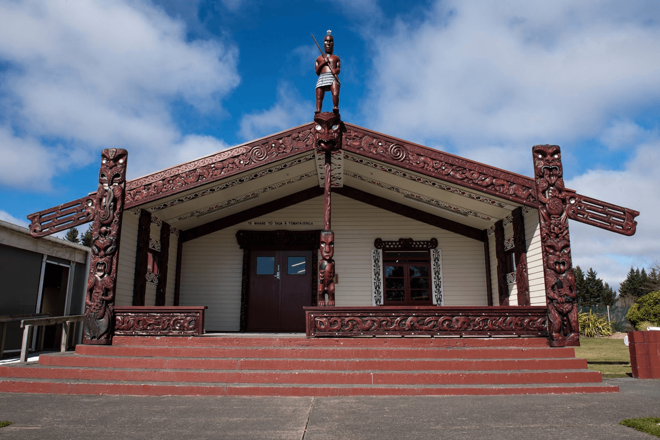 A traditional Māori meeting house with intricate red carvings and a statue on top. The building has a peaked roof with ornamental details and sits on a raised platform with steps. The sky is partly cloudy.