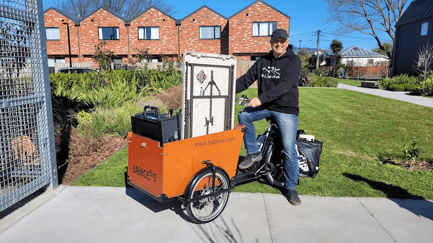 a white man with jeans smiling on a cargo bike with lots of stuff in the cart
