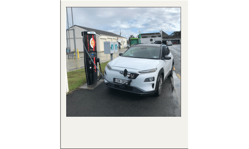 A white EV is parked outside in an area that looks like a petrol station. it is hooked up to a tall, black rectangular charging station.