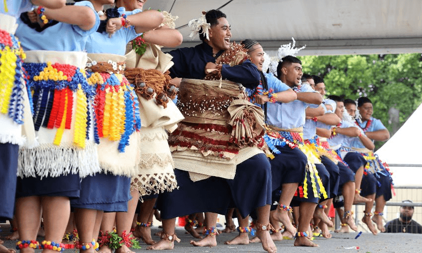 A group of people wearing colourful traditional Pacific Island attire performs a dance on stage. They are smiling, with festively decorated clothing, and adorned with flowers and woven accessories. Trees are visible in the background.