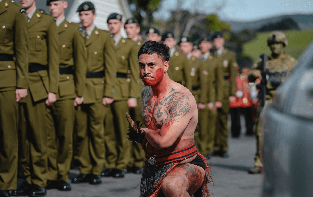A man performs a wero, wearing traditional attire with red body paint and intricate tattoos. He crouches on a street surrounded by uniformed soldiers standing in formation. The background features trees and a cloudy sky.