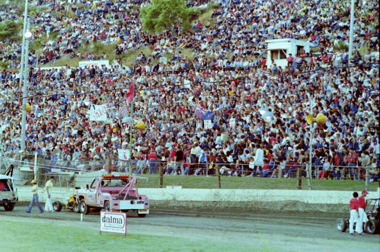 Crowd filling a large stadium for a motorsport event. A pink tow truck with a sponsor sign is on a dirt track. Many spectators are sitting on a grassy hill, and some are holding signs or flags.
