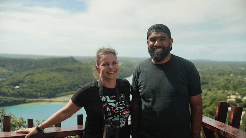 Nicola Toki and Pax Assadi stand together in front of a wooden fence. Behind them is a vast tropical landscape of sea and forest